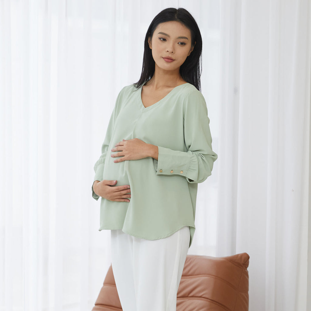 Pregnant woman in a light green blouse with white pants standing in a modern living room with large windows and natural light.