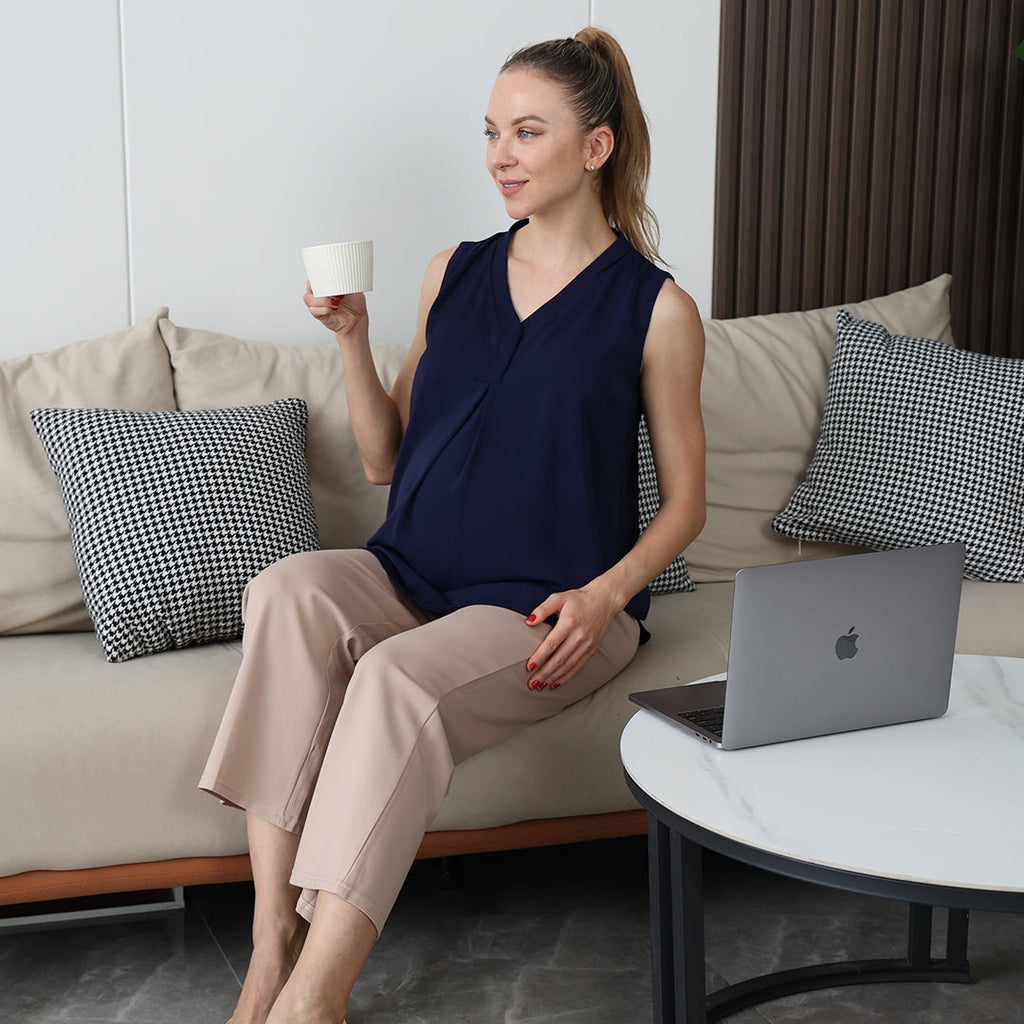 Pregnant woman in a navy blouse sitting on a sofa, holding a coffee mug, with a laptop on the table nearby.