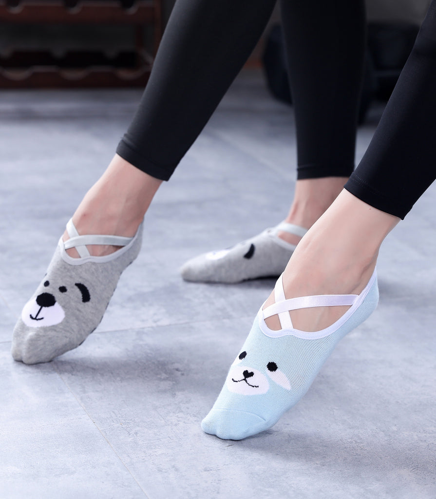 Women's feet in playful animal design socks, featuring a grey bear face on the left sock and a blue bear face on the right sock, resting on a light grey tiled floor.