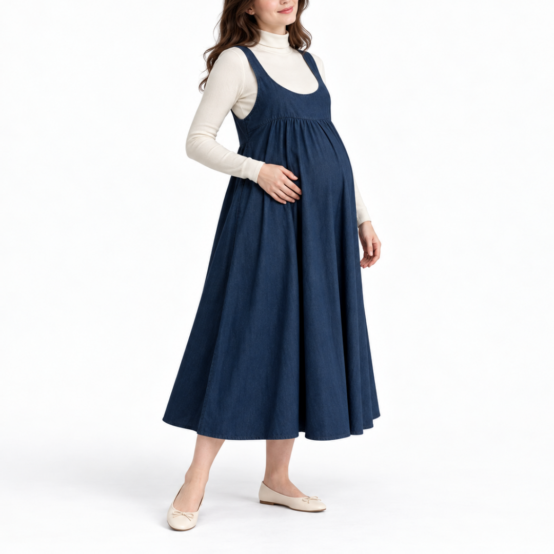 Angled front view of a pregnant model wearing a Blue sleeveless maternity pinafore dress over a white long-sleeve top.