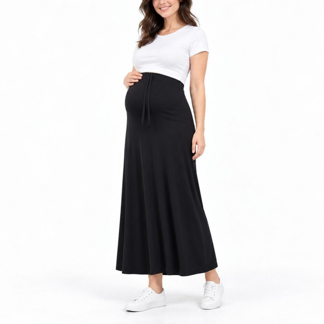 A pregnant woman modeling a high-waisted black maternity maxi skirt with a drawstring waist, paired with a white crop top and white sneakers, against a plain white background.