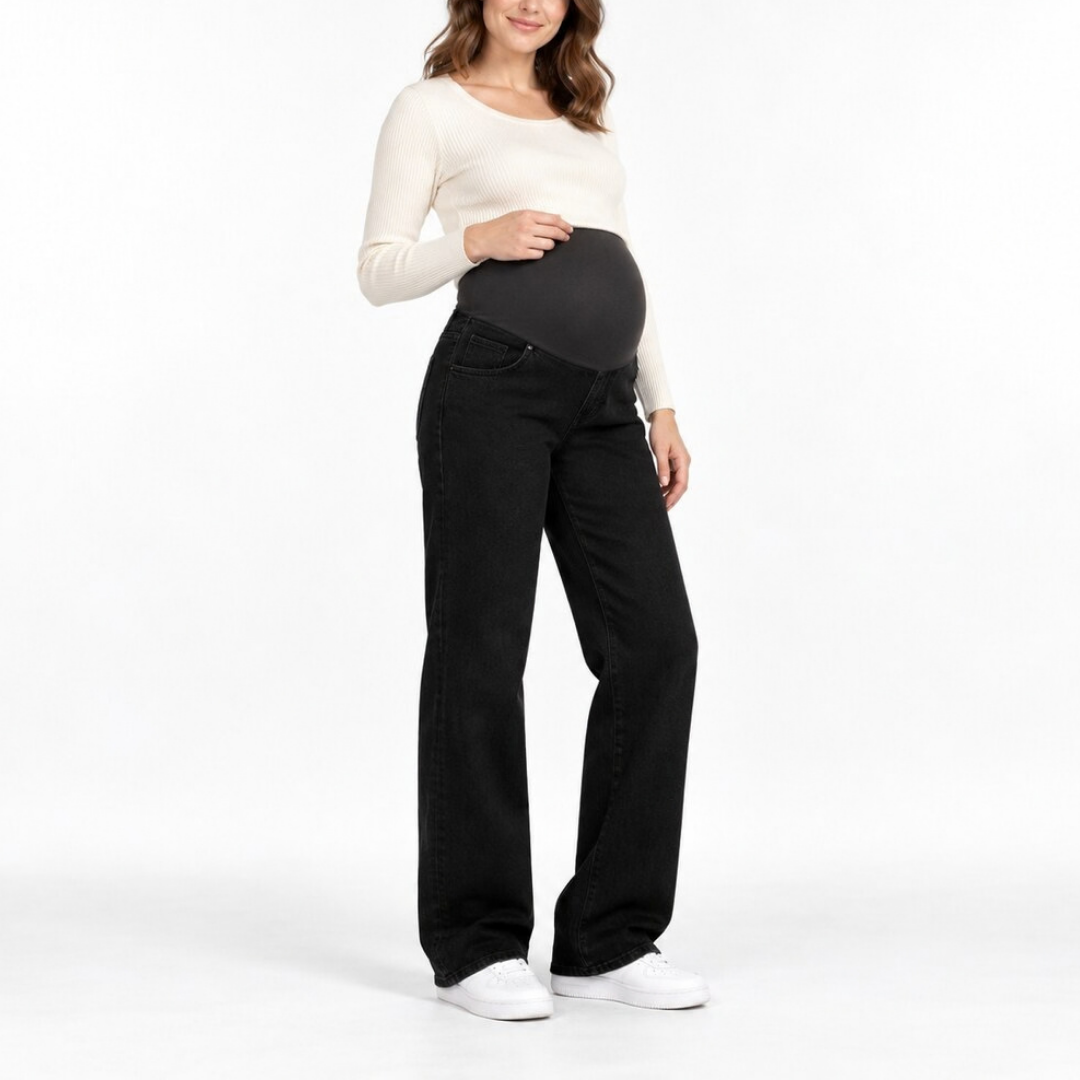 A pregnant woman wearing a black maternity belly support band over black jeans and a cream long-sleeve top, standing against a white studio background.