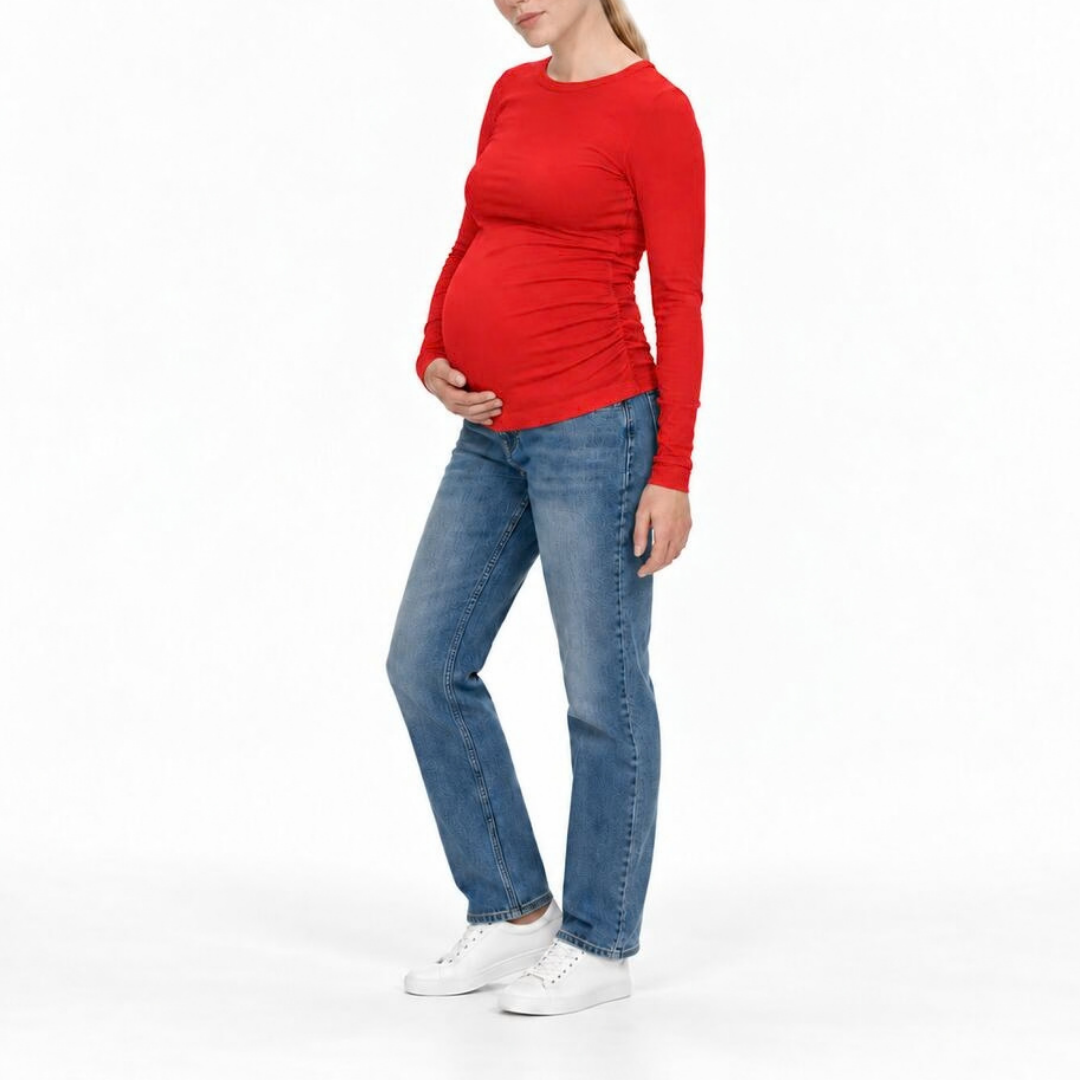 A pregnant woman wearing a bright red long-sleeve maternity top, blue jeans, and white sneakers, standing against a plain white studio background.