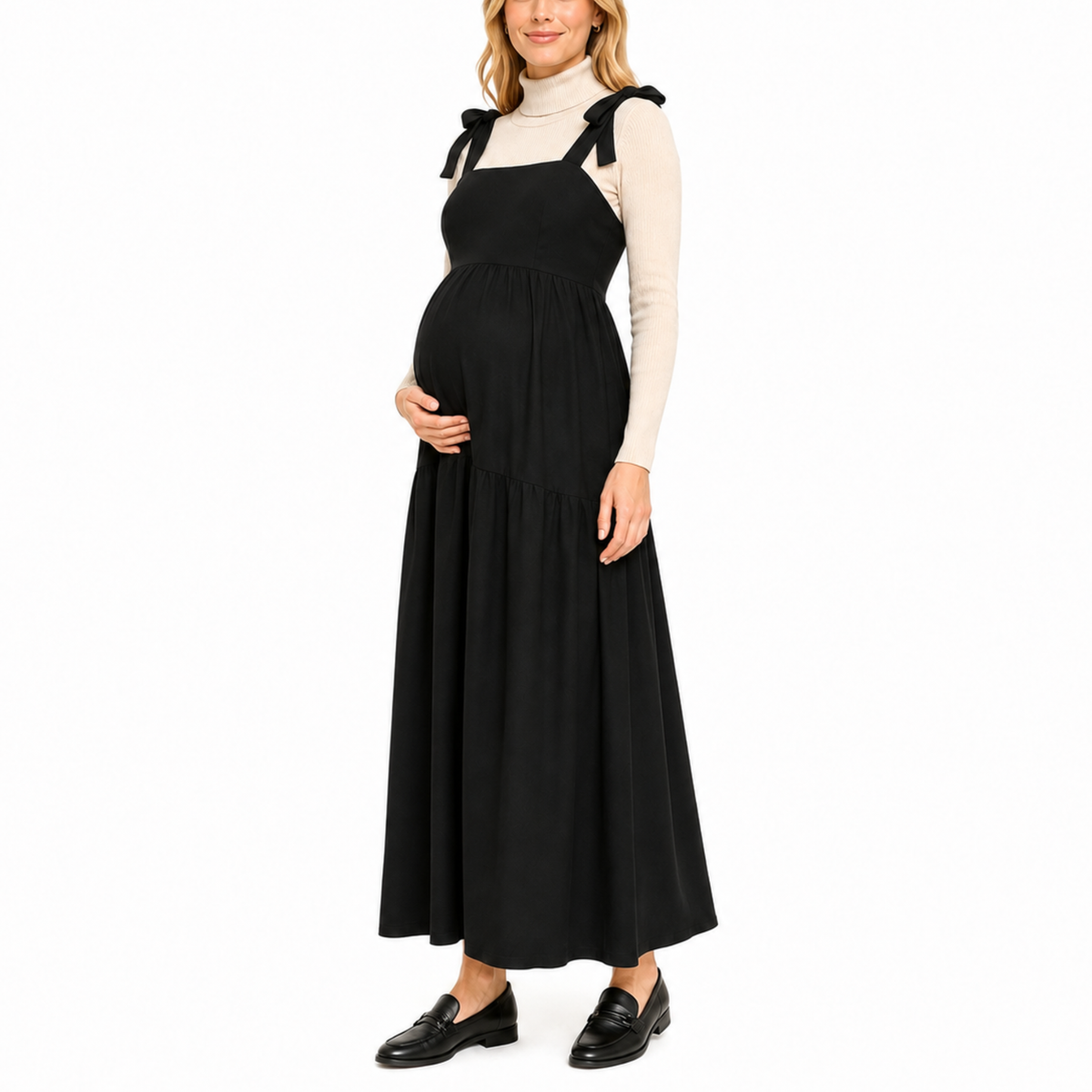 Pregnant model wearing a black solid maxi dress with a shirred bodice, tied shoulder straps, and a long flowing skirt layered over a white long-sleeve top.